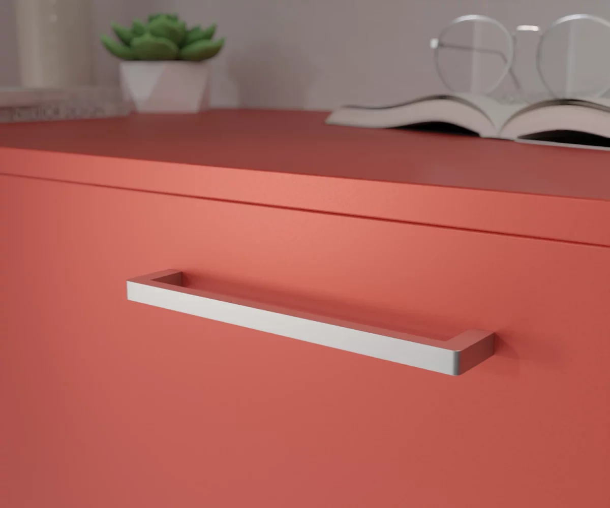 Silberfarbener Aluminium-Möbelgriff HA-AD-10004 mit streng rechteckigem Design und kantiger Silhouette. Der Griff besteht aus einem geraden, flachen Bügel mit gleichmäßiger Breite und rechtwinkligen Abschlüssen. Die Kanten sind präzise gearbeitet und die Vorderseite zeigt keinerlei Wölbung oder Rundung. Der Griff ist mittig auf einer korallenroten Schublade montiert dargestellt. Im Hintergrund unscharf dargestellt, befinden sich auf der rechten Seite ein aufgeschlagenes Buch mit einer Brille, die auf diesem Buch liegt. Auf der linken Seite befindet sich eine kleine grüne Pflanze in einem kleinen weißen Topf, links daneben eine hohe Vase und vor dieser Vase liegen zwei Bücher.
