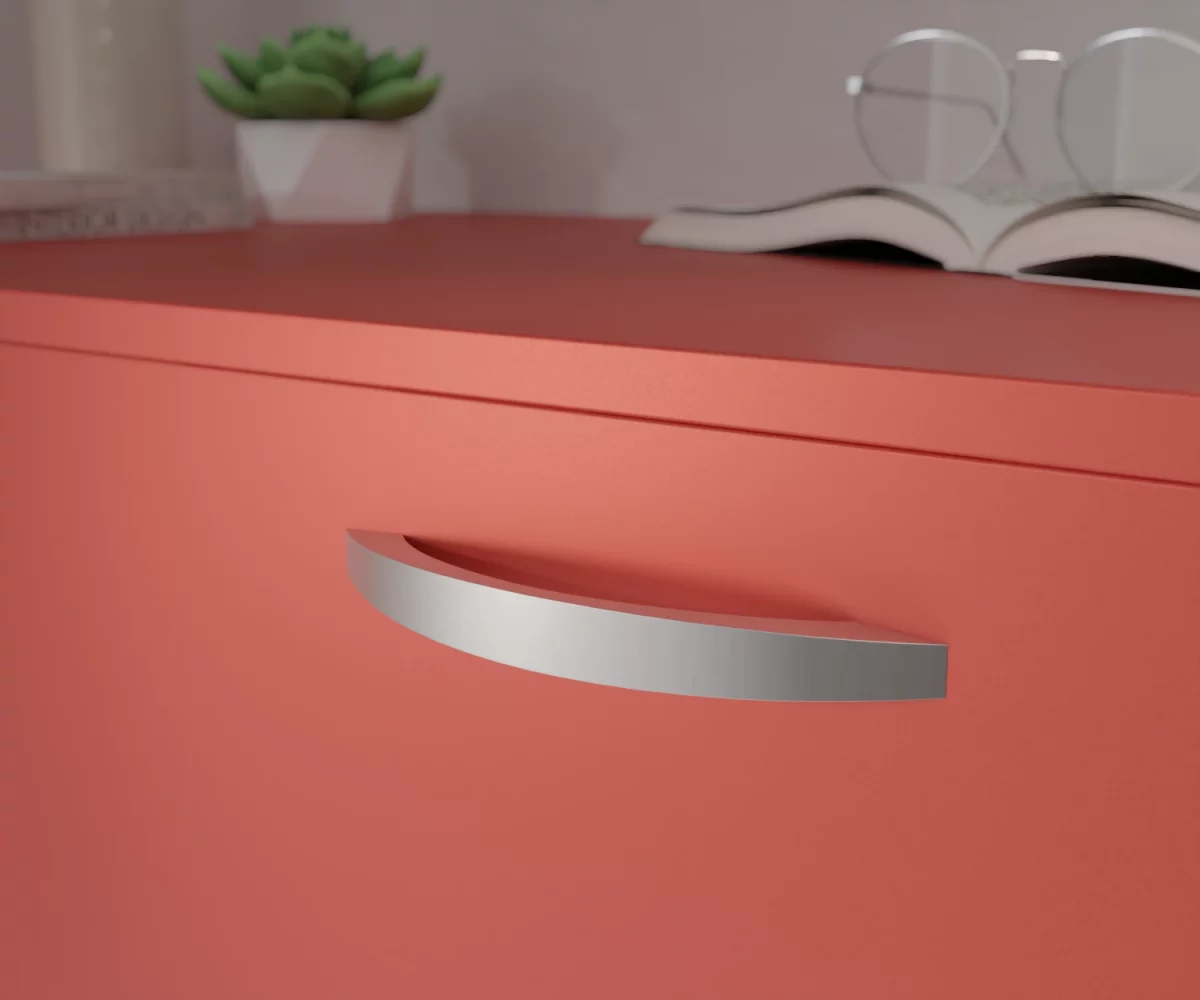 Silberfarbener Aluminium-Möbelgriff HA-AD-10019 mit gebogener Rückseite und leicht konvex gewölbter Vorderseite. Die Griffenden sind gerade geschnitten, die Seitenflächen verlaufen schmal und flach. Der Griff ist mittig auf einer korallenroten Schublade montiert dargestellt. Im Hintergrund unscharf dargestellt, befinden sich auf der rechten Seite ein aufgeschlagenes Buch mit einer Brille, die auf diesem Buch liegt. Auf der linken Seite befindet sich eine kleine grüne Pflanze in einem kleinen weißen Topf, links daneben eine hohe Vase und vor dieser Vase liegen zwei Bücher.