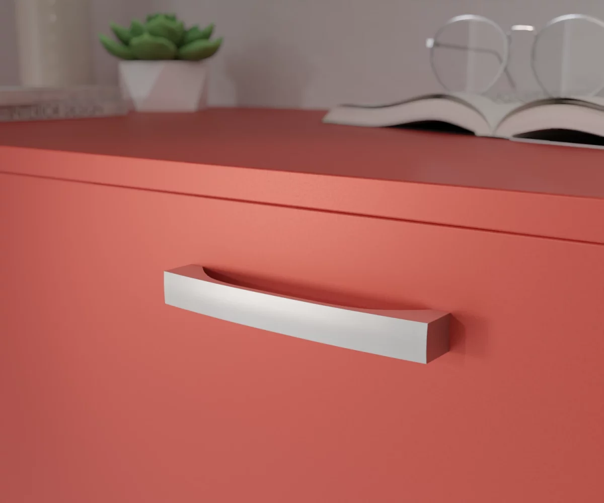 Silberfarbener Aluminium-Möbelgriff HA-AD-10021 mit geschwungener, konkaver Form, die sich zur Mitte hin nach innen wölbt und an den Außenseiten ausläuft. Die flachen, breiten Auflageflächen gehen fließend in die bogenförmige Griffpartie über. Der Griff ist mittig auf einer korallenroten Schublade montiert dargestellt. Im Hintergrund unscharf dargestellt, befinden sich auf der rechten Seite ein aufgeschlagenes Buch mit einer Brille, die auf diesem Buch liegt. Auf der linken Seite befindet sich eine kleine grüne Pflanze in einem kleinen weißen Topf, links daneben eine hohe Vase und vor dieser Vase liegen zwei Bücher.